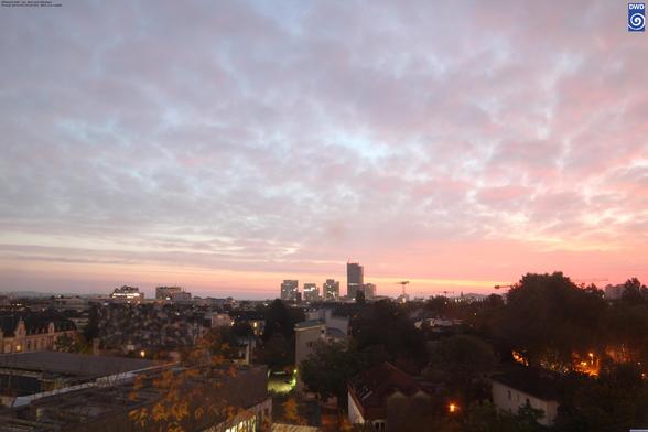 von der aufgehenden Sonne rötlich-gelblich angestrahlte flache tiefe Wolkenfetzen (Stratus), darunter die Lichter und Gebäude der Stadt Offenbach