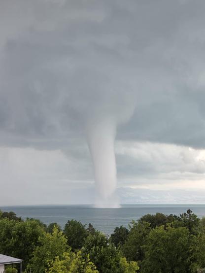 Große Wasserhose unter dunklen Gewitterwolken, im Vordergrund grüne Bäume und Uferlandschaft. Quelle: Nutzermeldungen DWD WarnWetter-App