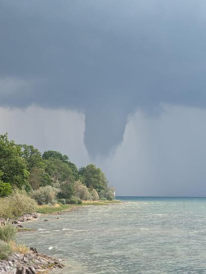 Dunkler Wolkentrichter über einem See, der sich nahe einer bewaldeten Uferlinie mit kleinem Leuchtturm absenkt. Quelle: Nutzermeldungen DWD WarnWetter-App