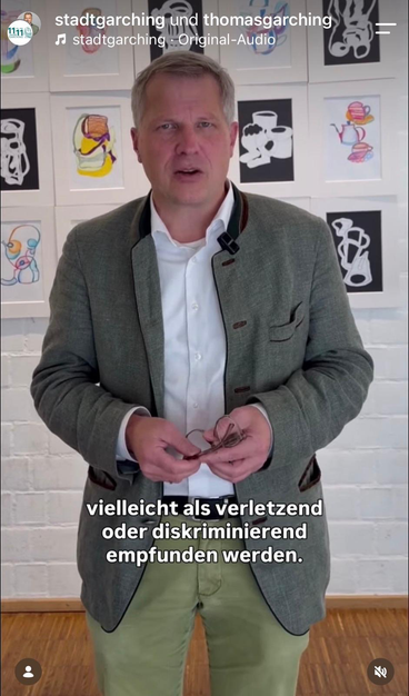 Bürgermeister Lemke steht vor einer Wand mit Kinderzeichnungen und hält eine Brille in den Händen. Die Untertitel lauten: „vielleicht als verletzend oder diskriminierend empfunden werden.“ Oben ist ein Instagram-Reel von @stadtgarching und thomasgarching zu sehen.