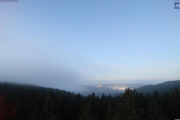 Wetterkamera Schmücke mit Blickrichtung Südwest. Im Tal beleuchtete Siedlungen, darüber ein Sternenhimmel, der von den ersten Strahlen der aufgehenden Sonne erhellt wird. Nebel- und Hochnebelschwaden wabern ebenfalls durch das Bild.
