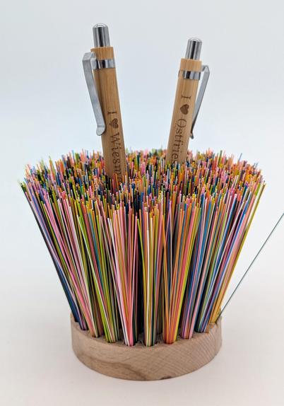 Ein Stifthalter, der aussieht wie eine runde Konfettibürste, die auf dem Rücken liegt und zwei Stifte (mit I love Ostfriesland und I love Wiesmoor) stecken zwischen den bunten Fasern