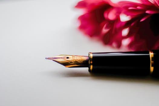 Nahaufnahme eines Füllhalters mit goldener Feder auf hellem Papier, im Hintergrund unscharfe pinke Blüte.