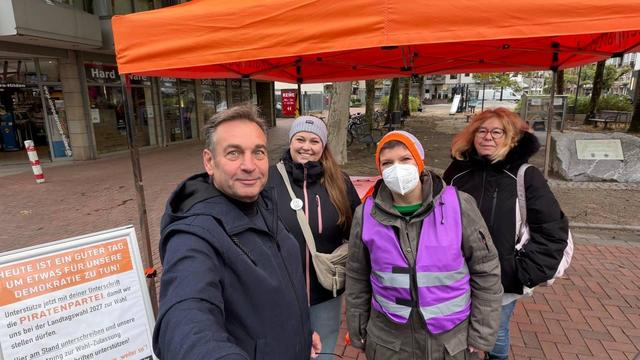 Selfie: Ralf, Anna, Jeanine und Gaby stehen unter einem orangefarbenen Pavillon. Links ein Kundenstopper mit Text für Unterstützungsunterschriften